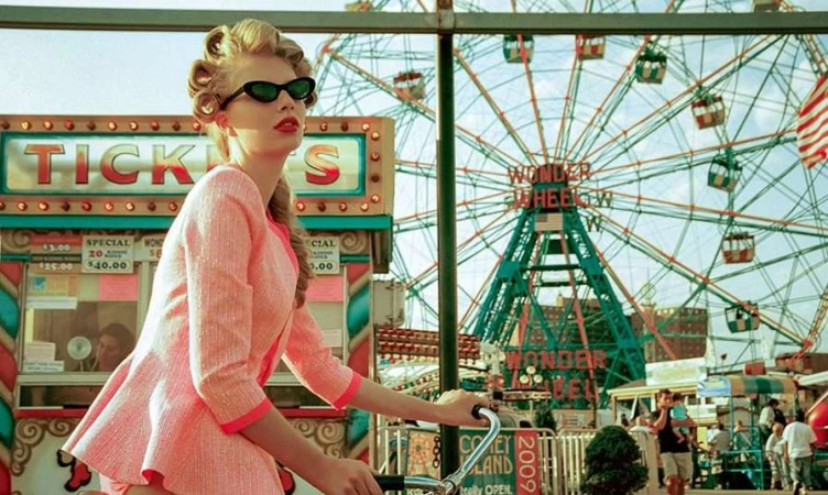 Blonde woman in pink dress and sunglasses riding a bicycle in front of a colorful Ferris wheel and ticket booth at Coney Island.