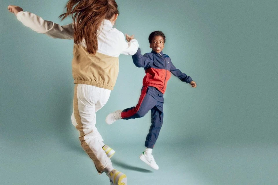 Two children jump in the air wearing color-blocked tracksuits against a pale blue background in a studio setting. 