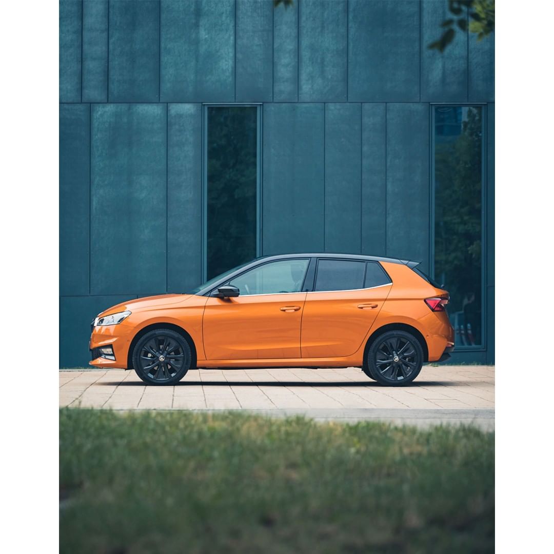 Orange Skoda Fabia hatchback parked on a paved surface in front of a modern, blue paneled building with green foliage visible.