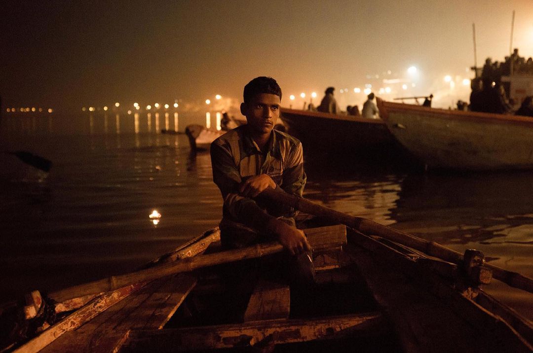 An adult male sits in a wooden rowboat on a body of water at night, with soft light reflecting off the water's surface and other boats visible.