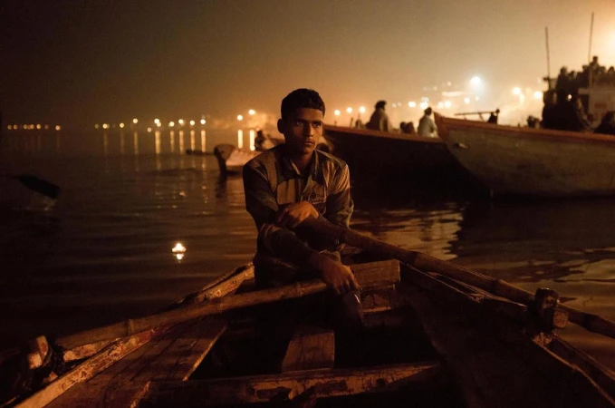 An adult male sits in a wooden rowboat on a body of water at night, with soft light reflecting off the water's surface and other boats visible.