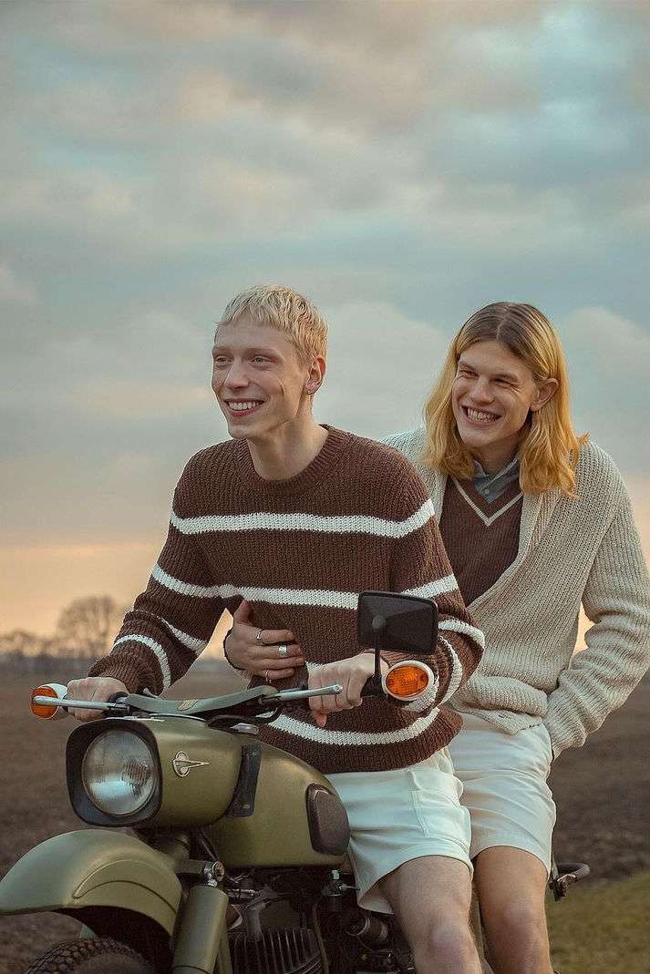 Two fair-skinned young men smile on an olive-green motorcycle against a cloudy sky, wearing knit sweaters and light-colored shorts.