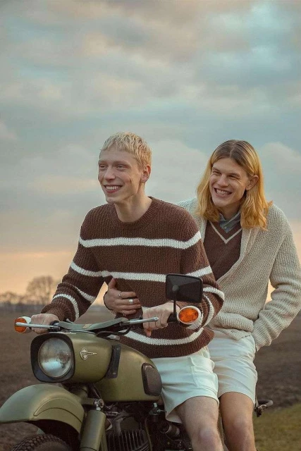 Two fair-skinned young men smile on an olive-green motorcycle against a cloudy sky, wearing knit sweaters and light-colored shorts.
