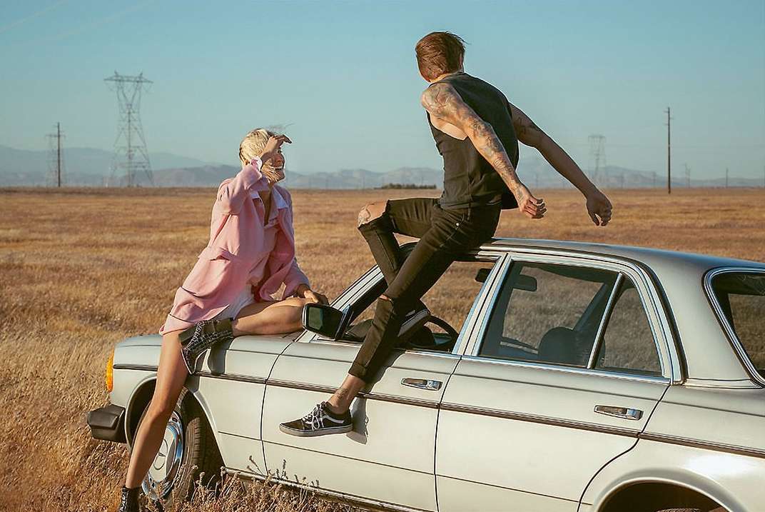Two models pose on a silver car in an open field under a clear sky, with power lines and distant mountains completing the scene.