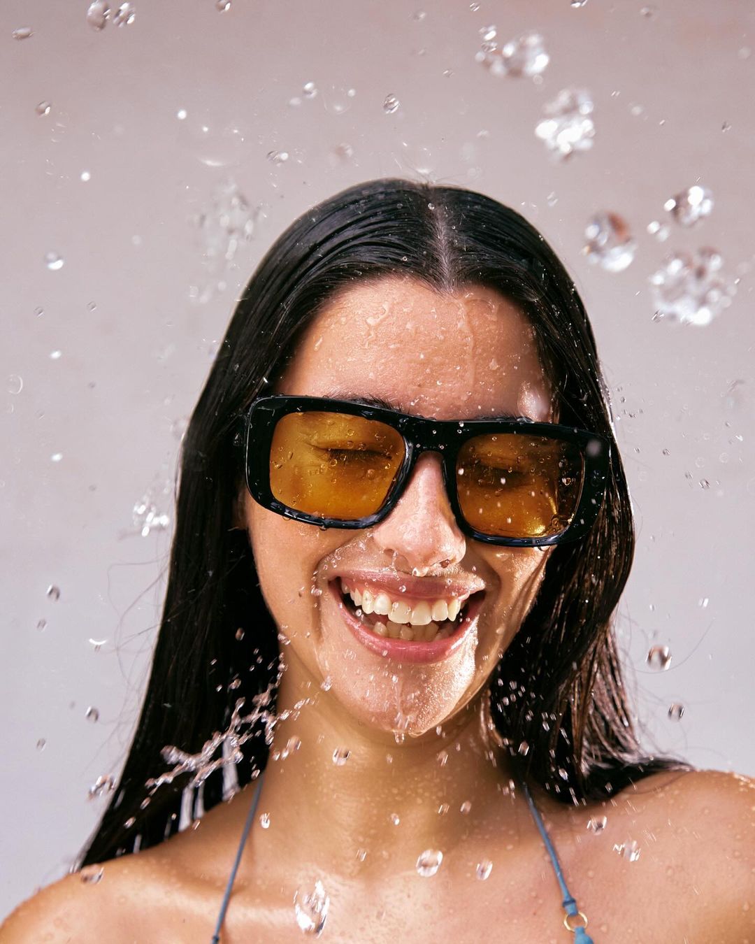 Portrait of a smiling woman with black hair wearing black-rimmed sunglasses with yellow lenses as water splashes against a light grey backdrop.