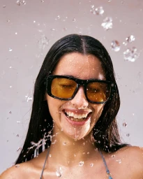 Smiling woman wearing black square sunglasses with yellow-tinted lenses as water splashes around her in a studio setting.