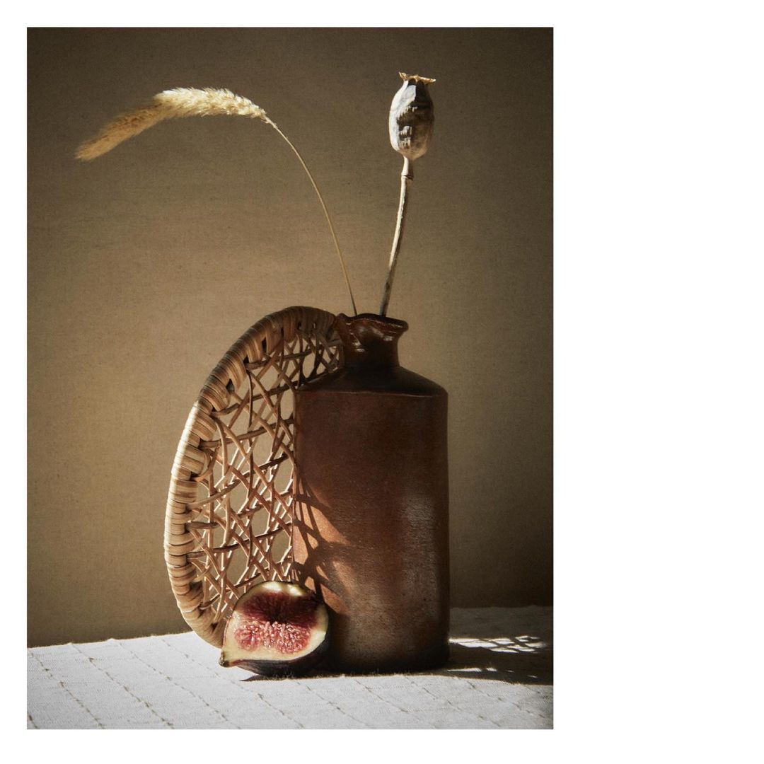 Still life photography featuring a vase with dried flowers, accompanied by a wicker plate and fig, set against a soft, warm-toned background.
