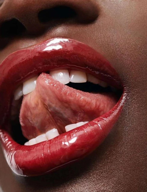 Close-up portrait of an African American adult female, showing lips covered in glossy red lipstick and tongue protruding between teeth.
