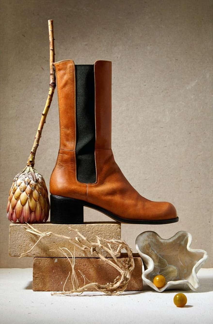 Studio shot of a tan leather boot on stacked bricks next to dried botanicals, a protea flower, and decorative glass dish.