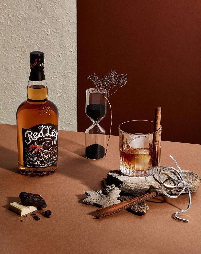 A still life showcases a liquor bottle, a faceted glass of liquor with ice, an hourglass, and chocolate against a neutral backdrop.