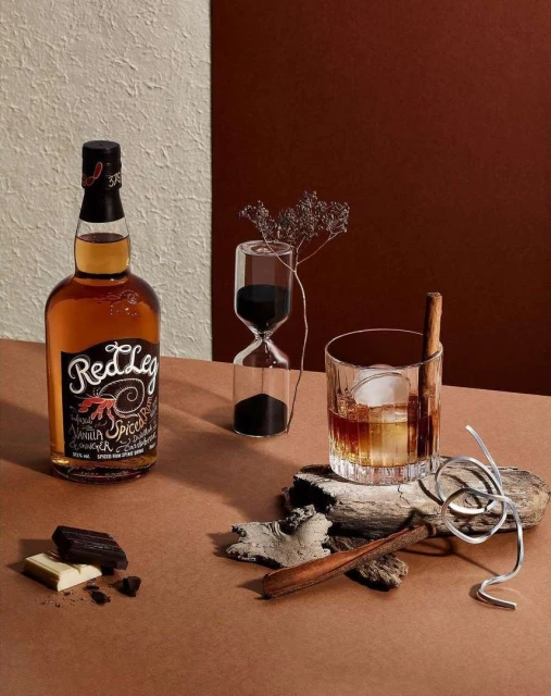 A still life showcases a liquor bottle, a faceted glass of liquor with ice, an hourglass, and chocolate against a neutral backdrop.