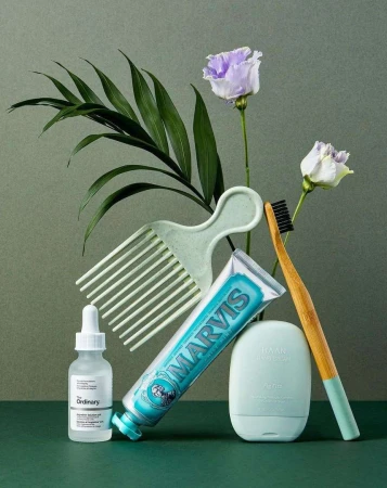Beauty and hygiene product still life featuring toothpaste, hair pick, toothbrush, dropper bottle, and flowers against green backdrop.