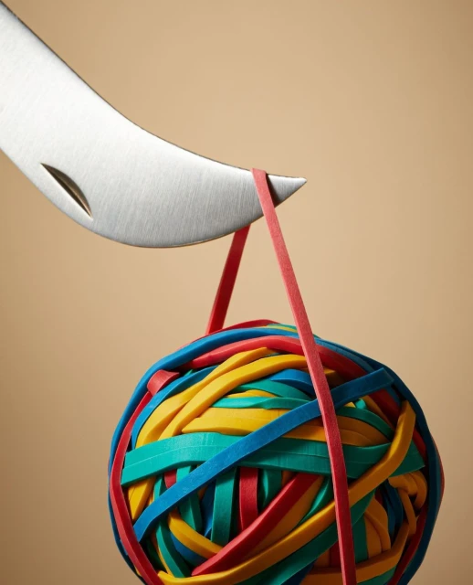 A silver knife slices a thick colorful ball of rubber bands with a red band acting as a hanger against a beige background.