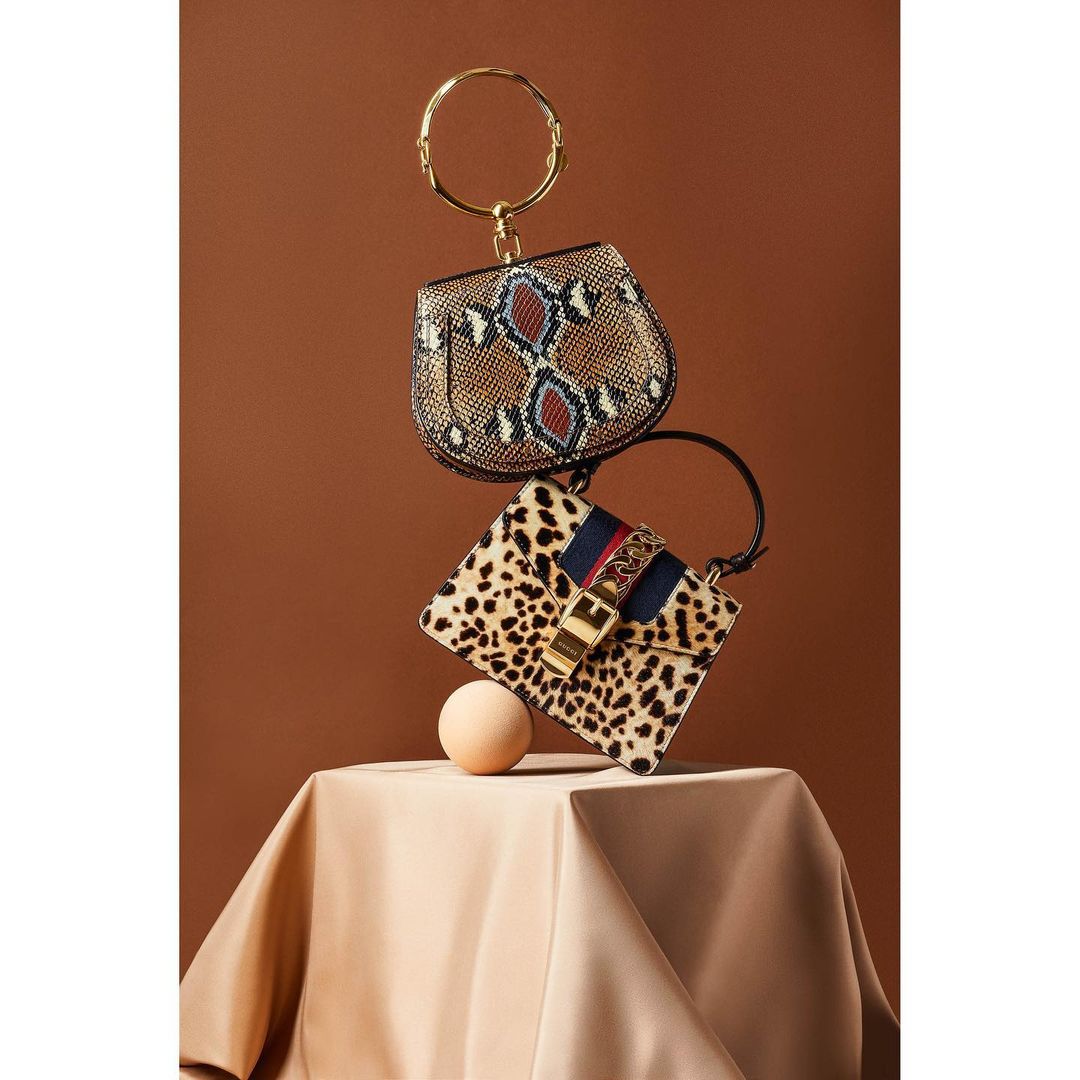 Still life studio shot of two fashion handbags in a stacked arrangement on a draped surface, with a seamless brown backdrop.