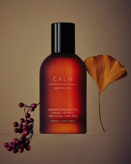 Calm room and linen spray in amber bottle with black cap, styled with a ginkgo leaf and red berries on a neutral surface.