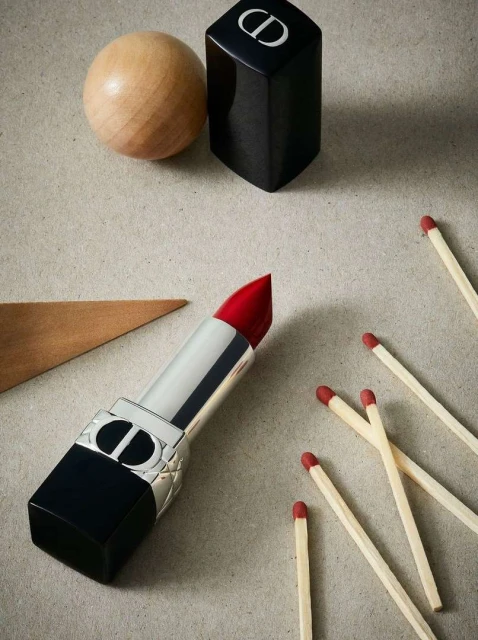 Still life of Dior lipstick with wooden ball, triangle and matches on a textured surface. Cosmetics and geometric shapes.