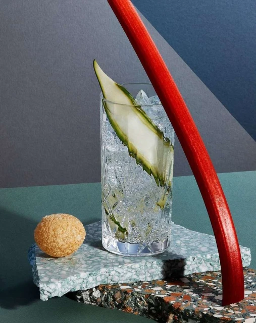 Still life of a cocktail in a patterned glass with a cucumber garnish and a rhubarb stalk, set on terrazzo platforms against a gray background.