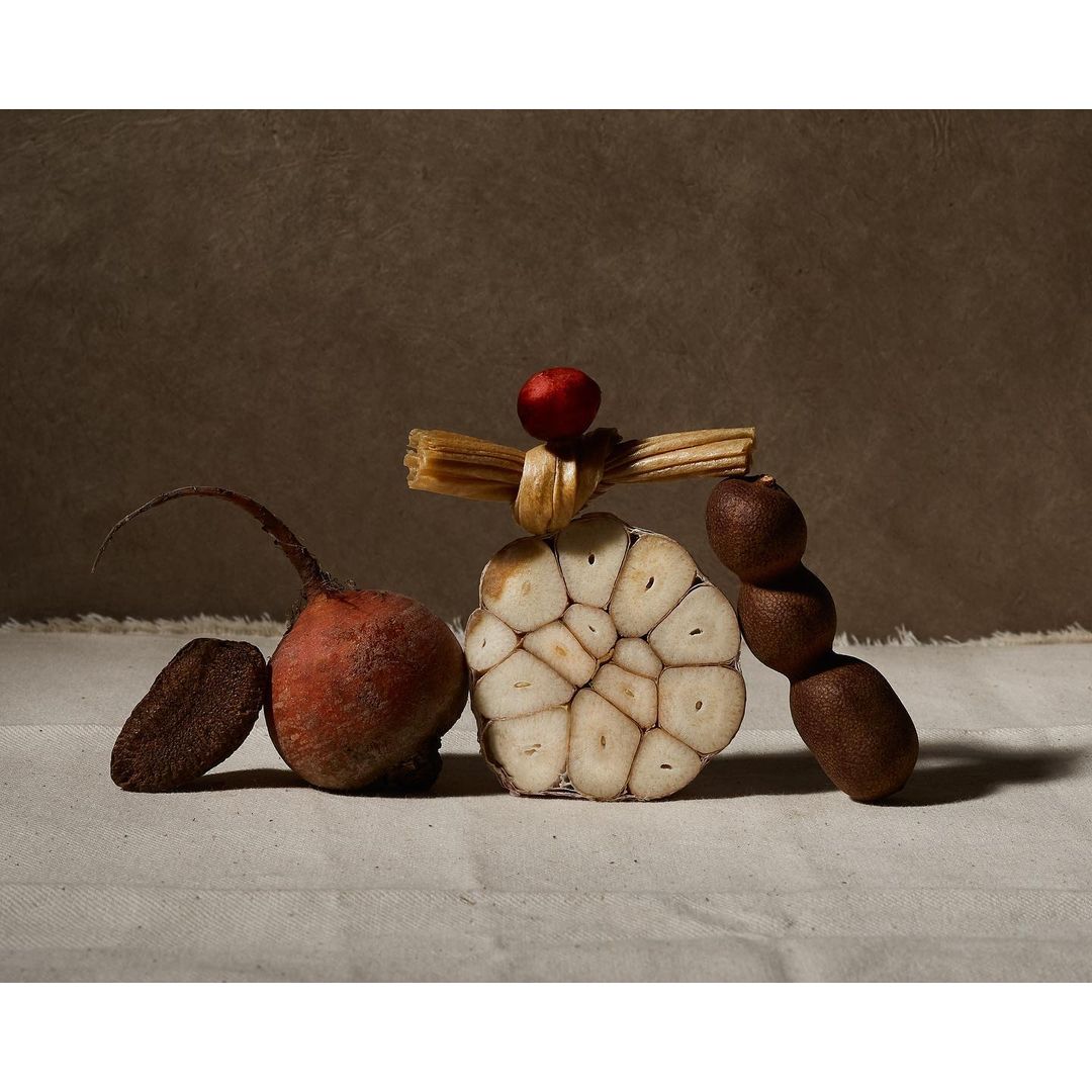 Still life photograph of a beet, garlic, small red fruit and a stack of wheat on a linen cloth against a brown background.
