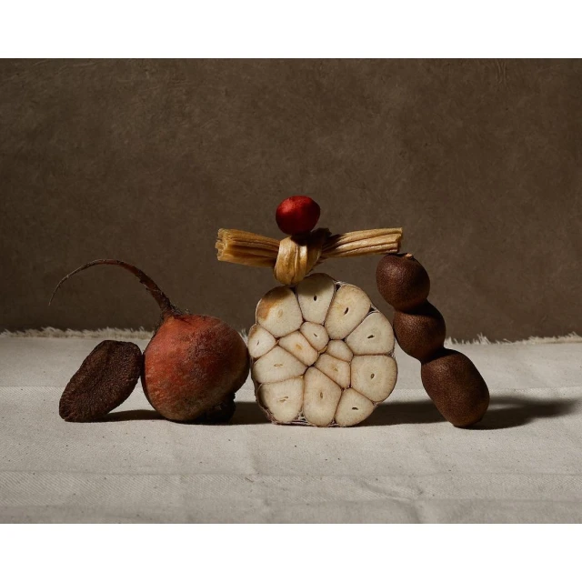 Still life photograph of a beet, garlic, small red fruit and a stack of wheat on a linen cloth against a brown background.