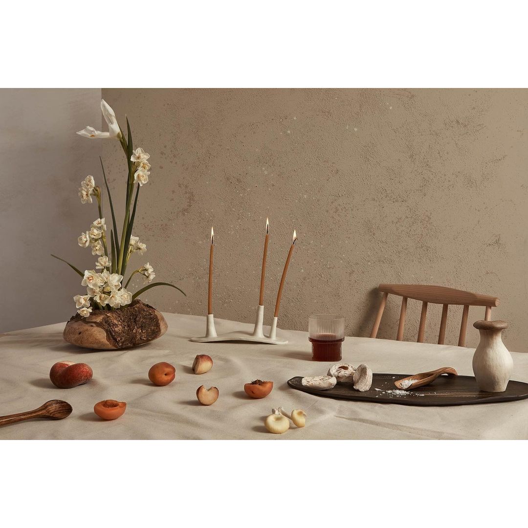 Still life of peaches, flowers, and candles arranged on a linen tablecloth in a natural light setting. Neutral color palette and textures.