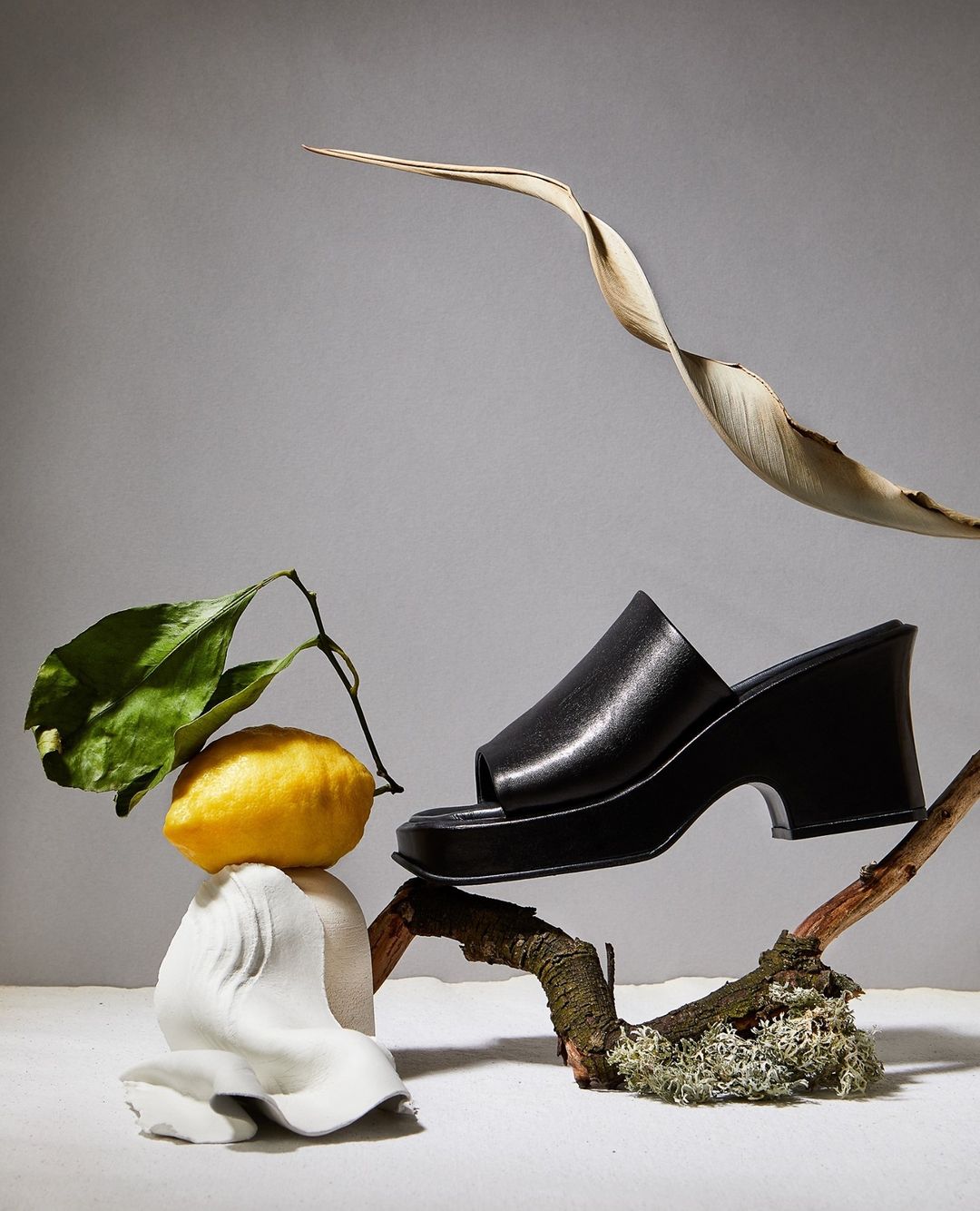 Still life fashion product shot of a black platform sandal displayed with lemon, leaves, and sculpted rock against a neutral backdrop.