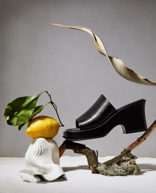 Still life fashion product shot of a black platform sandal displayed with lemon, leaves, and sculpted rock against a neutral backdrop.