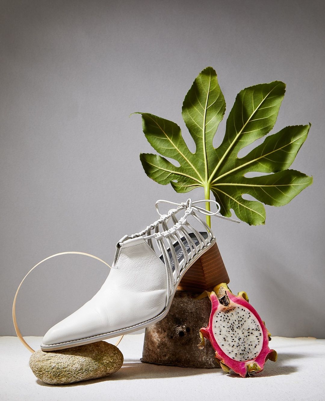 Studio still life of a white ankle boot with decorative straps, dragon fruit, and large leaf against a pale grey backdrop.