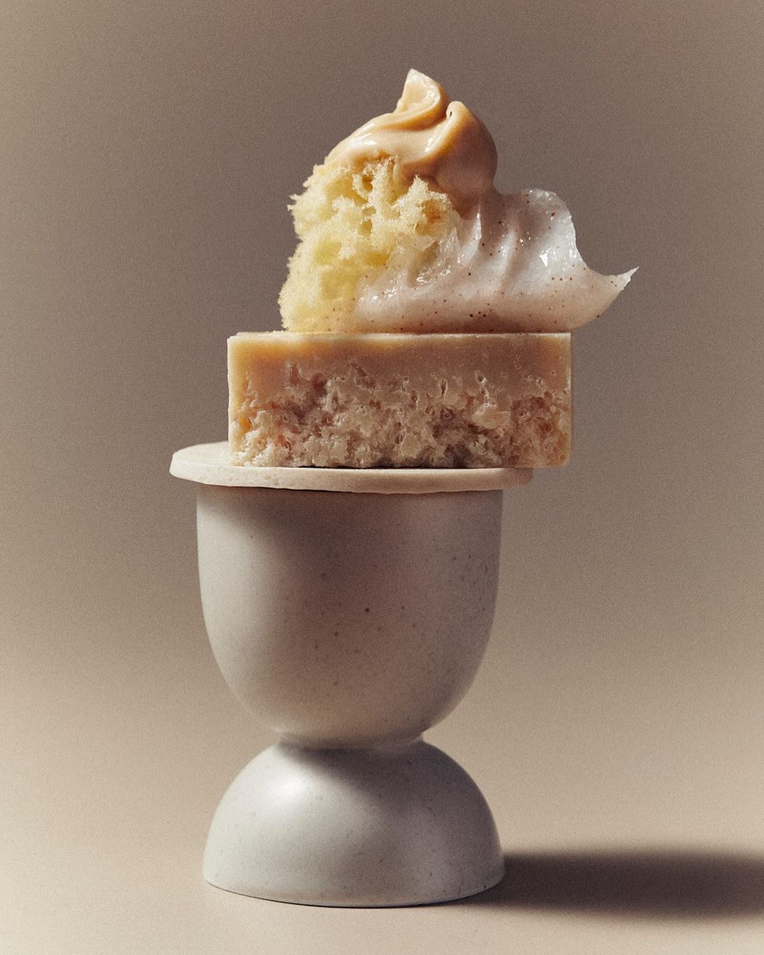 Stacked cubes of cake and icing on a pedestal, set against a soft-beige background for a minimalist food or dessert presentation.