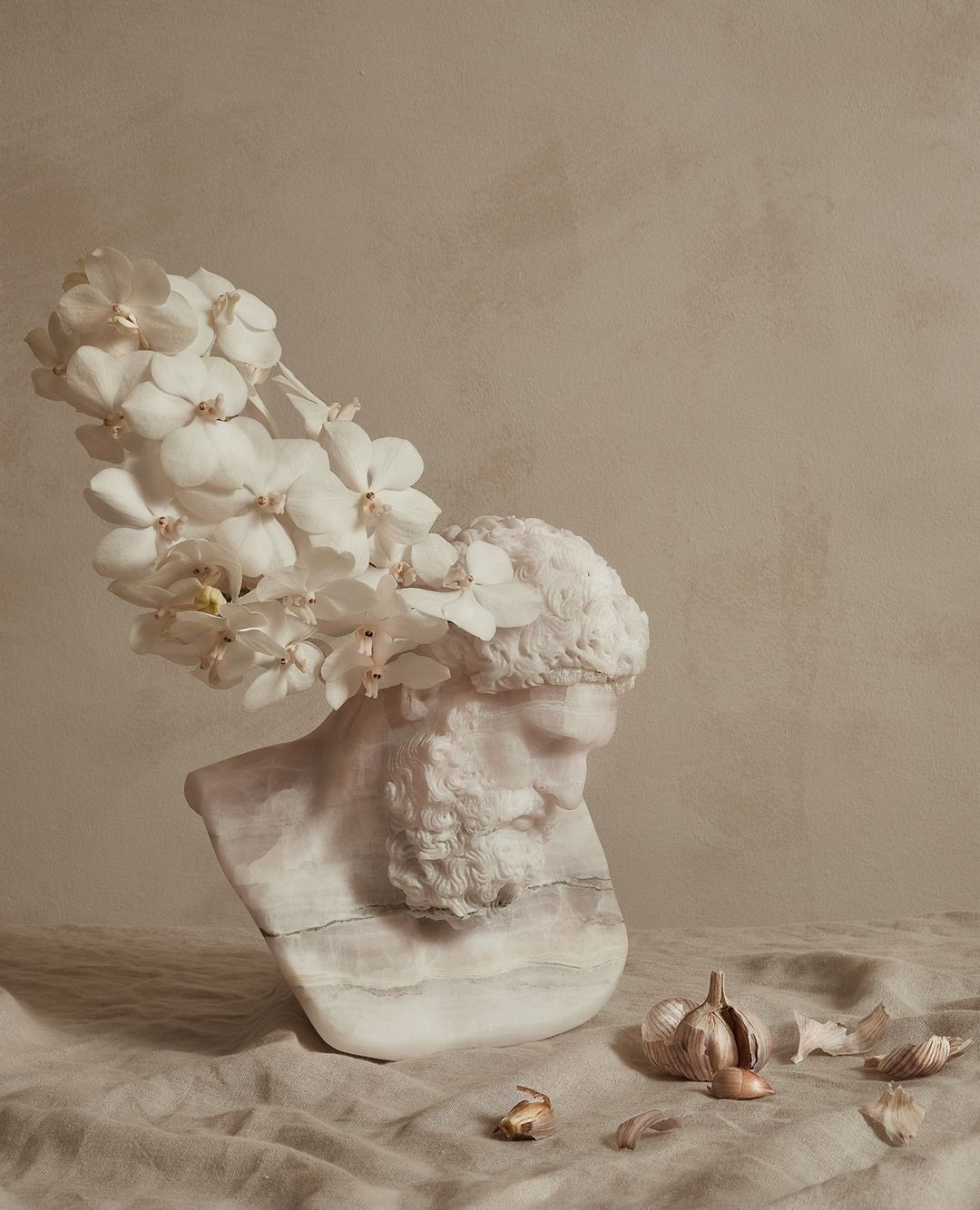 Bust sculpture of a bearded man with white orchids on his head, alongside garlic bulbs and cloves on a linen surface in a studio.