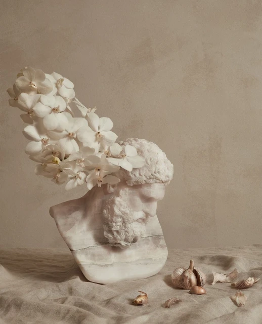 Bust sculpture of a bearded man with white orchids on his head, alongside garlic bulbs and cloves on a linen surface in a studio.
