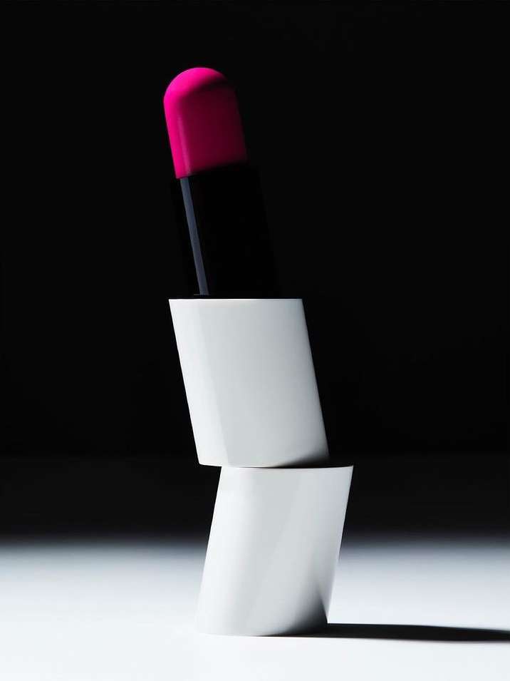 Close-up product photography of a bright pink lipstick standing on stacked white geometric cylindrical tubes against a dark background.
