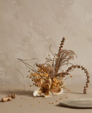 Still life featuring a lily, daisies, and dried grasses in a vase, complemented by scattered seeds and nuts against a neutral background.