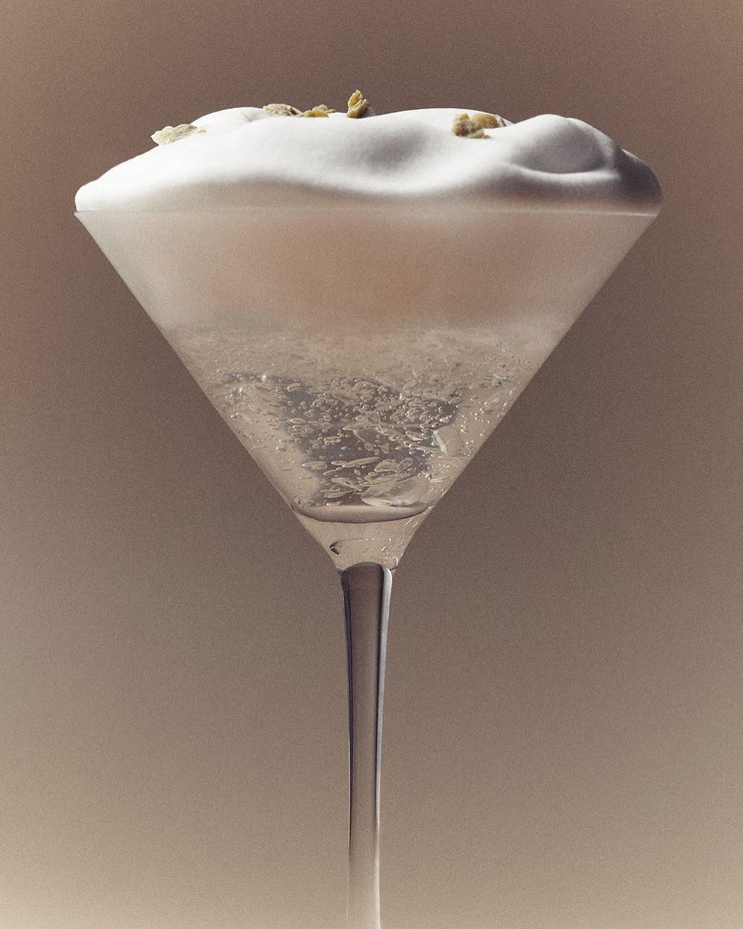 Close-up studio shot of a sparkling martini cocktail in a clear glass with white foam and garnish, on a plain beige background.