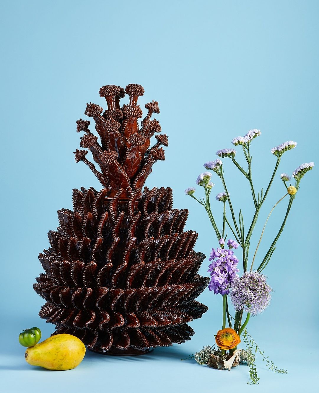 Still life photography with a large, decorative pinecone-shaped object alongside purple and white flowers, a yellow pear, and a lime.