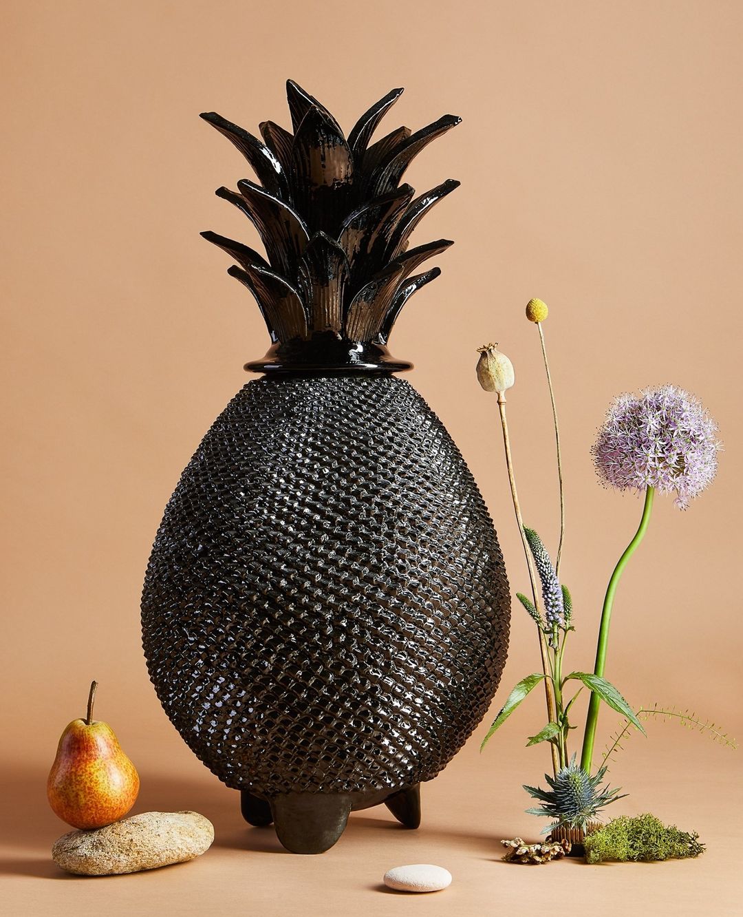 Still life of a black pineapple-shaped lamp, pear, and wild flowers including thistle and allium on a peach colored background.