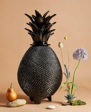Still life of a black pineapple-shaped lamp, pear, and wild flowers including thistle and allium on a peach colored background.