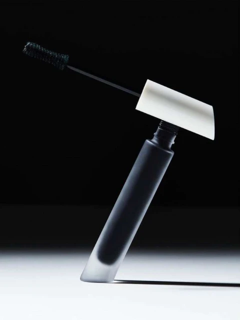 Studio shot of a black mascara tube with a white applicator, angled against a black background with bright white light and shadow.