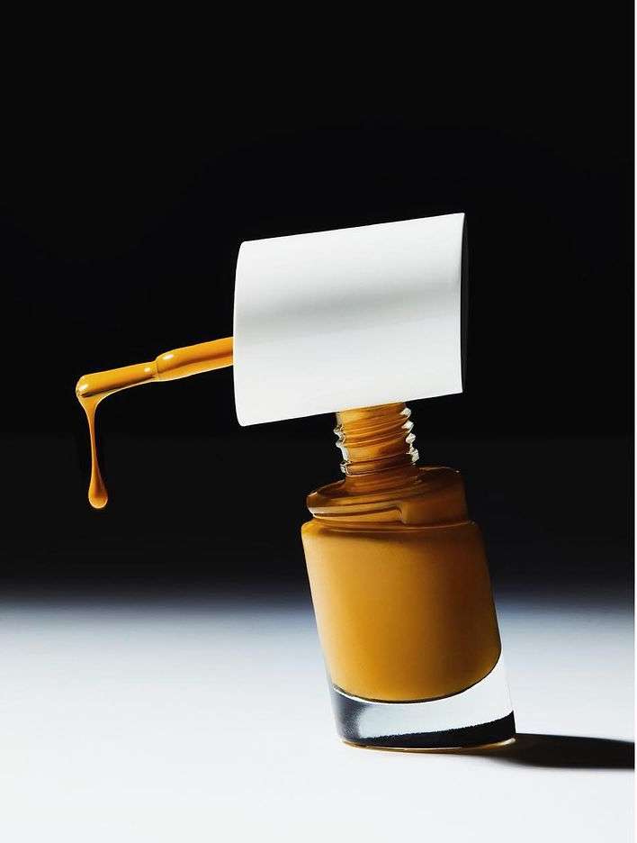 Close-up studio shot of an open bottle of caramel nail polish with the applicator brush resting on the bottle, dripping a drop of polish.