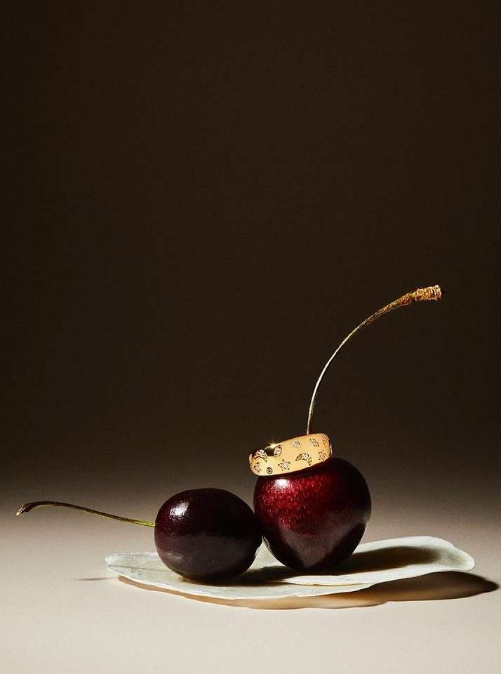 Studio shot of two cherries and a golden ring with sun and moon designs. Dark background, moody lighting, jewelry product shot.