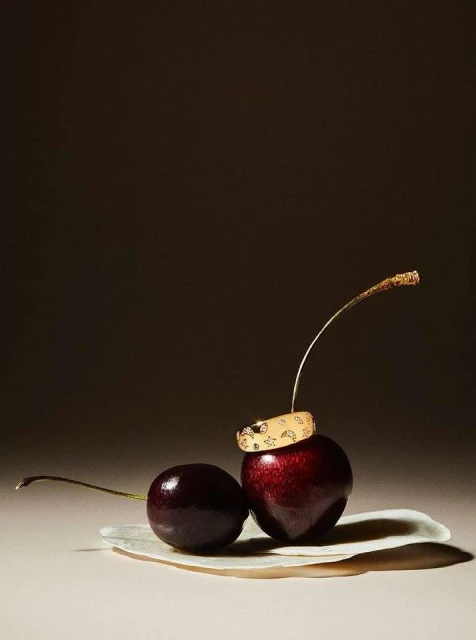Studio shot of two cherries and a golden ring with sun and moon designs. Dark background, moody lighting, jewelry product shot.
