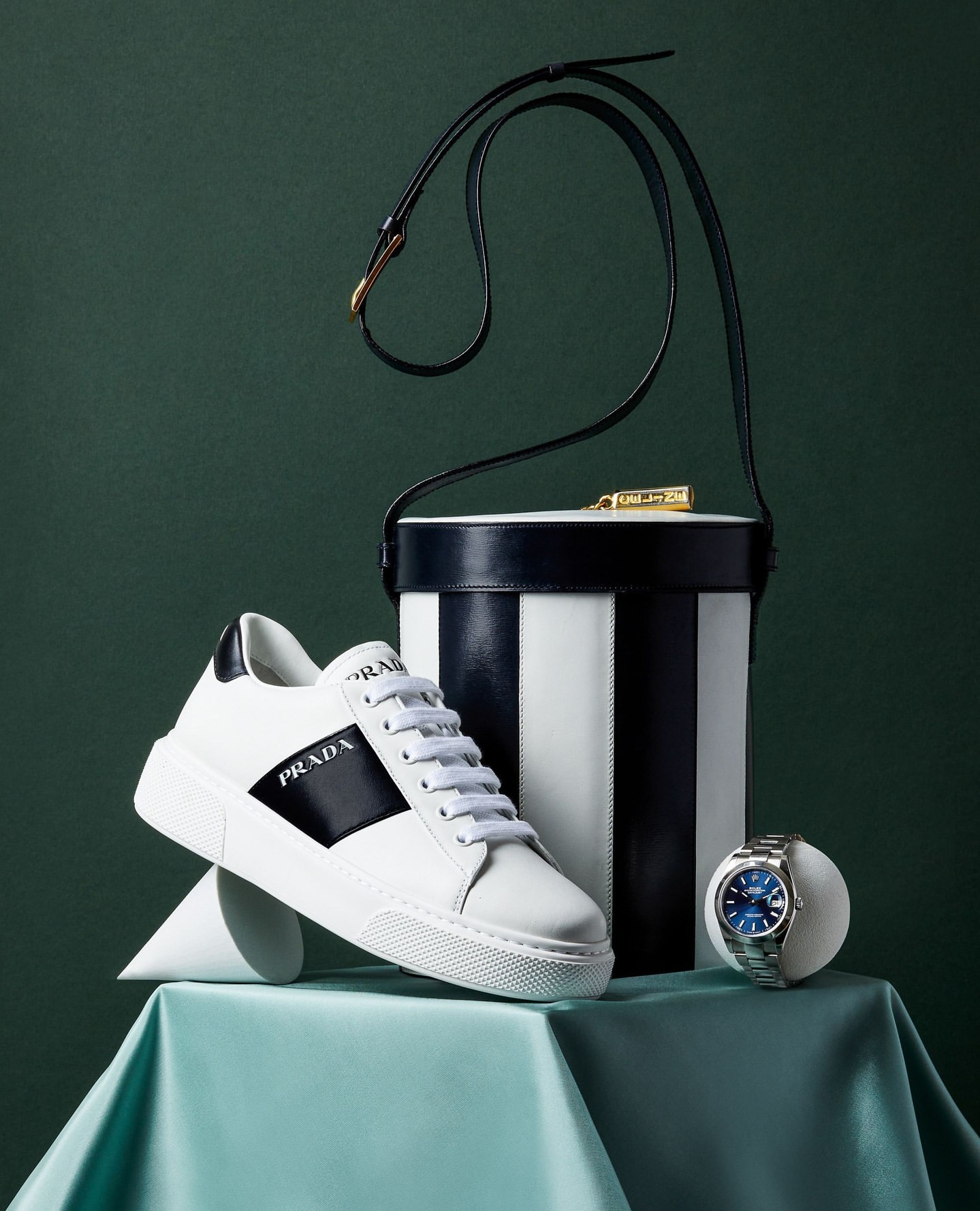 Elevated studio shot of a Prada sneaker, striped handbag, and blue-faced watch, set against a green backdrop, showcasing luxury fashion.