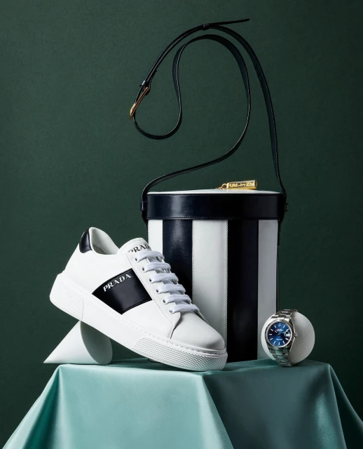 Elevated studio shot of a Prada sneaker, striped handbag, and blue-faced watch, set against a green backdrop, showcasing luxury fashion.