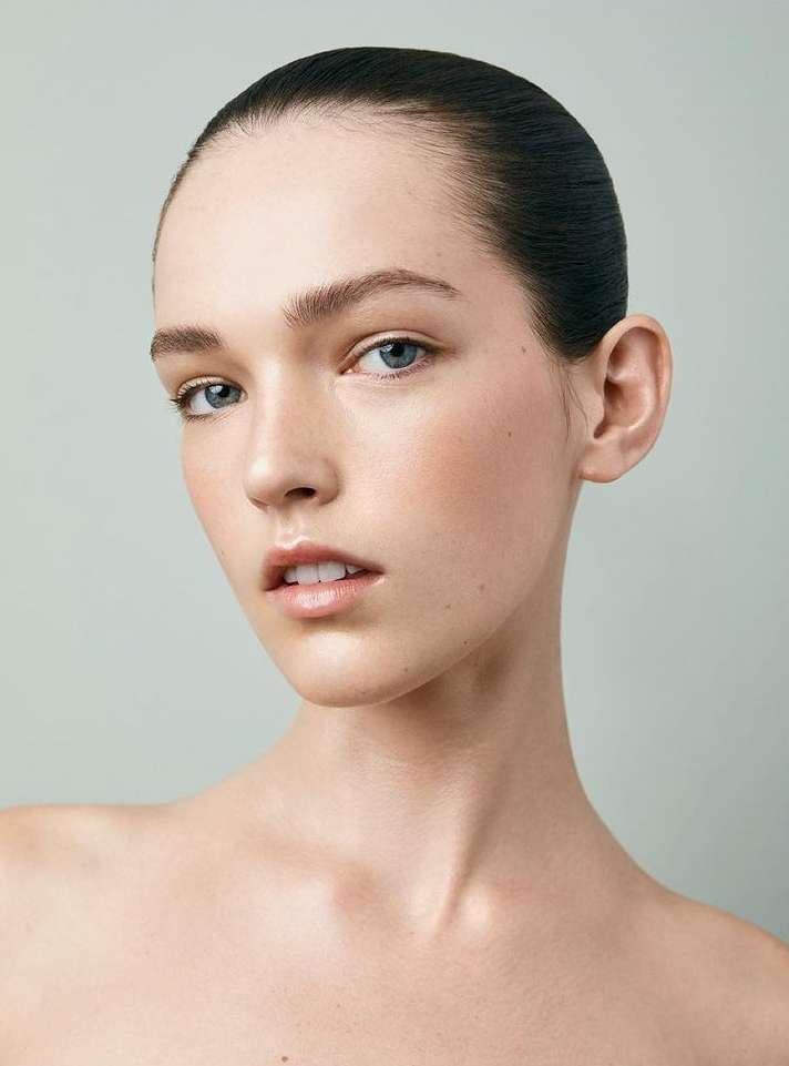 Close-up portrait of a young woman with slicked-back dark hair, fair skin, and light eyes against a light grey studio background. Beauty shot.