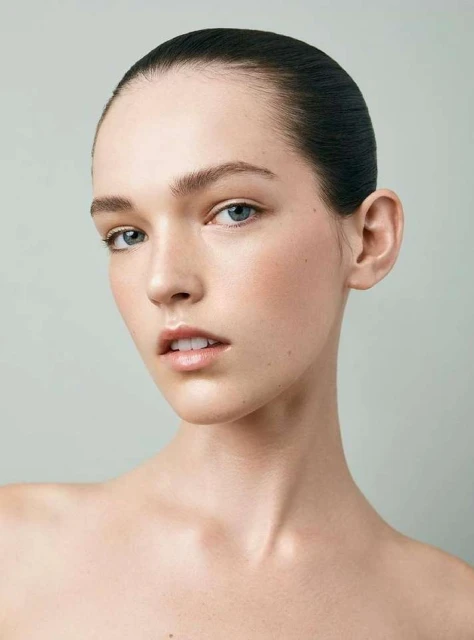Close-up portrait of a young woman with slicked-back dark hair, fair skin, and light eyes against a light grey studio background. Beauty shot.