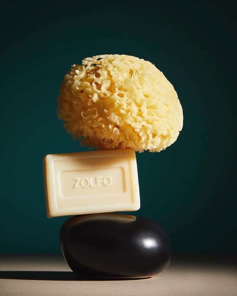 Still life studio shot of a natural sponge, bar of Zolfo soap, and black stone stacked against a teal backdrop with soft, diffused lighting.