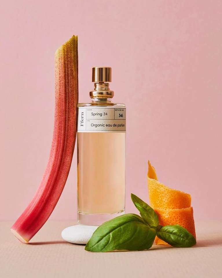 Still life of a perfume bottle, rhubarb stalk, basil leaves, orange peel curl, and white stone against a pink background.
