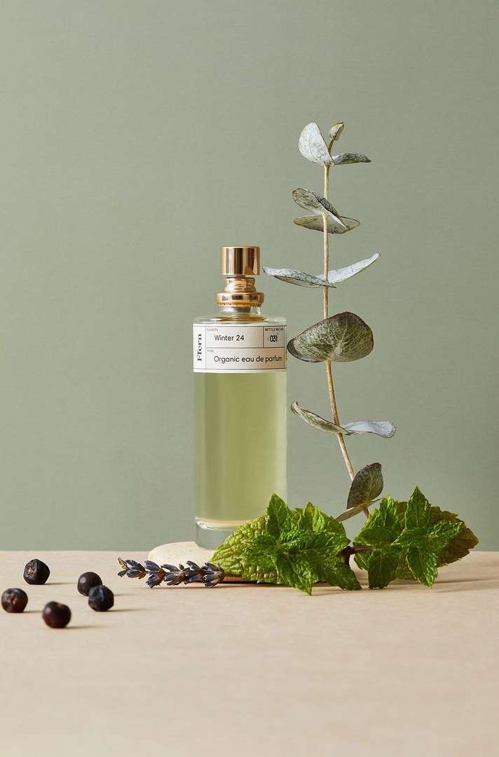Perfume bottle and botanicals, featuring a clear bottle, gold cap, green foliage, and dark berries against a muted green backdrop.