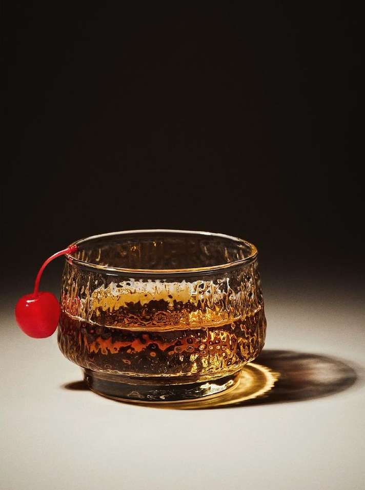 Short textured glass filled with amber liquid and a red maraschino cherry resting on the rim, set against a dark gradient background.