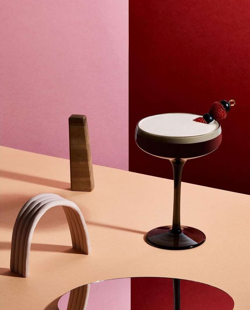 Elegant cocktail with foam and berry garnish on a peach surface with minimalist geometric shapes and a dual-tone pink and red background.