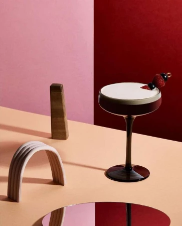 Elegant cocktail with foam and berry garnish on a peach surface with minimalist geometric shapes and a dual-tone pink and red background.